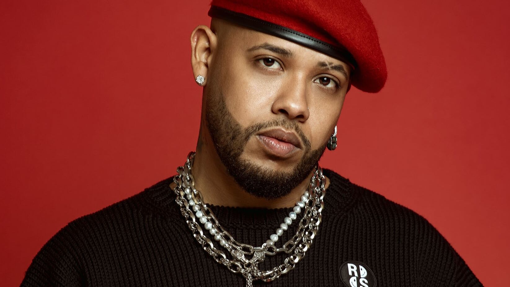 Rafa Pabón posa sobre un fondo rojo, con una camisa manga larga negra, prendas y un gorro rojo.