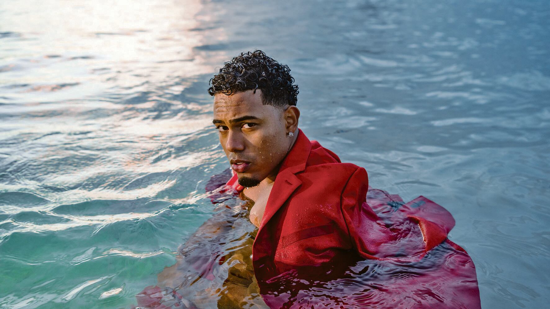 Myke Towers lleva puesto un traje rojo en una piscina.